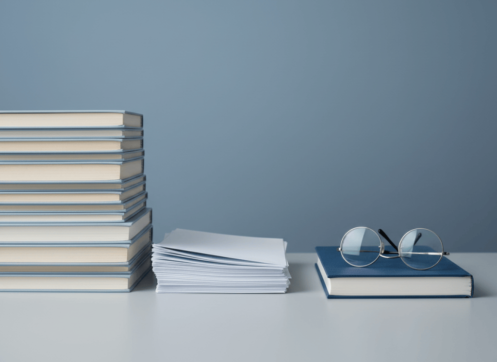 Minimalist academic still life representing literature reviews: neatly stacked medical research journals and papers on a light gray desk beside reading glasses, cool blue-gray palette, soft even lighting, clean editorial photography, no text.